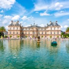 In front of the Luxembourg Palace lies a large lake with a scenic central fountain. The water reflects the elegant palace façade, surrounded by tulips and manicured gardens.