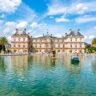 In front of the Luxembourg Palace lies a large lake with a scenic central fountain. The water reflects the elegant palace façade, surrounded by tulips and manicured gardens.