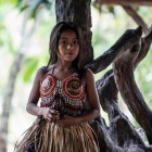 A young Wayuri indigenous girl, dressed in traditional attire, leaning against a tree in the Ecuadorian Amazon jungle.