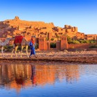 The fortified village of Ait Benhaddou, a UNESCO site in Morocco, is reflected in the water among camels, earthen walls and sunset light.