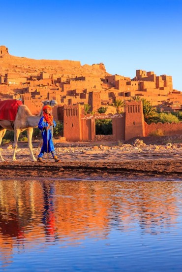 The fortified village of Ait Benhaddou, a UNESCO site in Morocco, is reflected in the water among camels, earthen walls and sunset light.