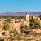 The view over Ouarzazate reveals ochre-colored buildings, historic kasbahs, and palm groves, with the High Atlas Mountains providing a striking backdrop.