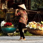 A Vietnamese vendor in Hanoi amidst historic streets and markets, with baskets of flowers bringing scents and colors to everyday city life.