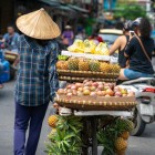 A street vendor on a bicycle carries tropical fruits through the streets of ancient Hanoi, among old houses and lively street life.