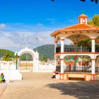 The square of Zinacantán, in front of the Church of San Lorenzo, features an elegant white-and-orange gazebo decorated with colorful banners, surrounded by green hills and authentic local life.