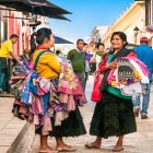 Two Tzotzil women walk through the streets of San Cristóbal de Las Casas carrying embroidered clothing and linens, offering local handicrafts and keeping Indigenous traditions alive.