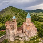Aerial view of Gremi fortress in Kakheti: stone towers and church with terracotta roofs dominate the hill amid green valleys and mountains of eastern Georgia.