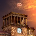 The clock tower of the Government Building in Republic Square, Yerevan, Armenia, glowing at sunset with the moon rising in the background sky.