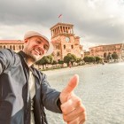 Smiling tourist taking a selfie in front of the fountains of Republic Square in Yerevan. In the background stands the Government Building clock tower and elegant Armenian architecture.
