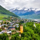 View of Mestia at dusk, with illuminated medieval towers among green valleys and traditional houses, framed by the majestic Caucasus mountains.