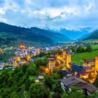 View of Mestia at dusk, with illuminated medieval towers among green valleys and traditional houses, framed by the majestic Caucasus mountains.