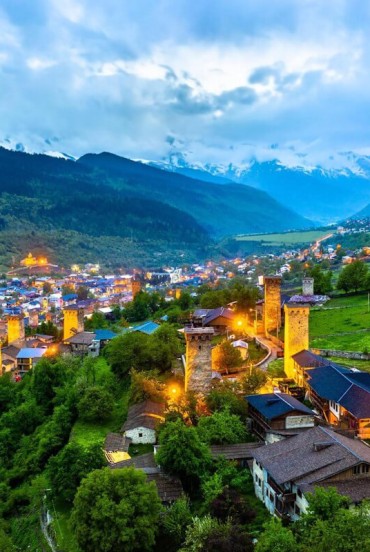View of Mestia at dusk, with illuminated medieval towers among green valleys and traditional houses, framed by the majestic Caucasus mountains.