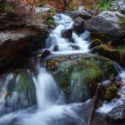 Gveleti waterfall in Dariali Gorge: crystal-clear water flows over mossy rocks and autumn leaves, creating a fresh and evocative atmosphere in the heart of Georgian nature.