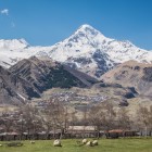 View of Gergeti village at the foot of Mount Kazbek, with houses gathered in the valley and the snow-capped Greater Caucasus peaks dominating the horizon.