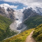 Panoramic trail through green meadows and mountain light, at the foot of the Shkhara glacier. The path leads the gaze toward the Caucasus peaks, between silence and open spaces.