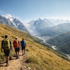 Three hikers walk along the Chkhunderi Pass trail, through golden meadows and high-altitude silence. Ahead, the valley opens toward the river and the snow-capped Caucasus peaks.