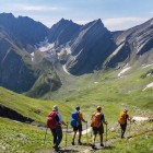Hikers walking along a trail in the Greater Caucasus, between green valleys and rocky ridges. The path descends through open silence, with lingering snow guiding each step.