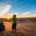 Sunrise among the dunes of Merzouga, with two travellers watching the sun rise over the Sahara, surrounded by silence, golden sand and endless horizons.