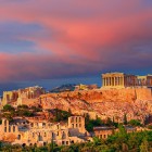 At sunset, the Acropolis rises above Athens with the Parthenon glowing in warm pink hues. A breathtaking panorama celebrating over two thousand years of Greek history.