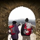Two pilgrims walk along Saint James Street in the village of Portomarín, where the road ahead continues along the Santiago Way, framed by an ancient stone arch.