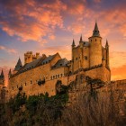 The fairytale silhouette of the Alcázar of Segovia crowns the rocky promontory, while the sunset sky bathes its towers, battlements, and walls in a golden light that enhances the elegance of its medieval architecture.