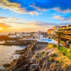 At sunset, the promenade of Puerto de Santiago follows the volcanic coastline among palm trees, terraces and pools overlooking the Atlantic, as the sky glows with warm golden hues.