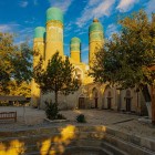 In the peaceful courtyard of the Chor Minor Madrasa, trees and arcades lead to four turquoise-domed towers, an unmistakable symbol of historic Bukhara.