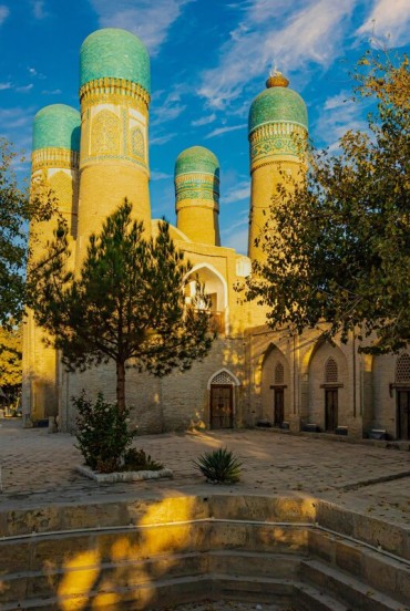 In the peaceful courtyard of the Chor Minor Madrasa, trees and arcades lead to four turquoise-domed towers, an unmistakable symbol of historic Bukhara.