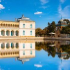 Beyond the entrance, the route reaches the palace gardens, where the pale façade and pavilion are reflected in the large pool, offering a first view of the entire complex.